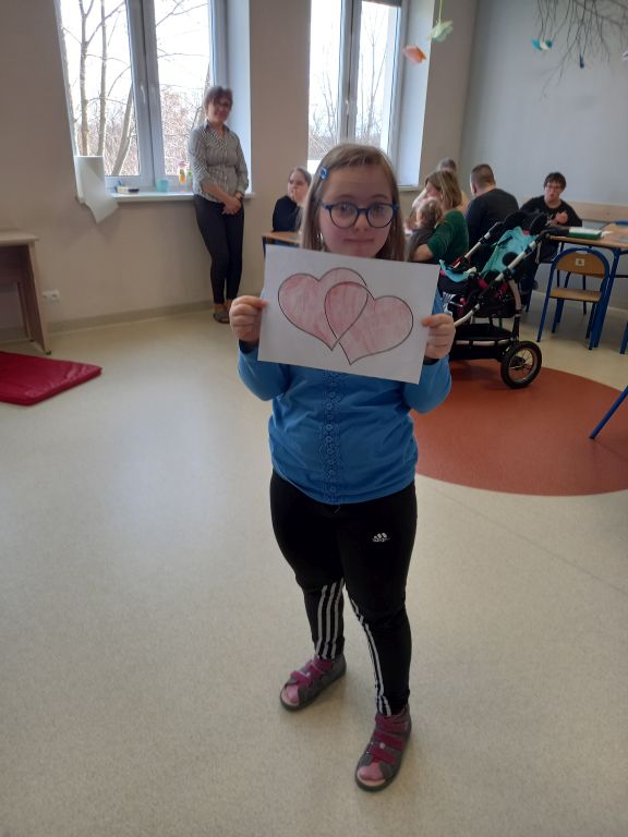 Fotografia przedstawia dziewczynkę prezentującą do zdjęcia swoją pracę plastyczną w postaci narysowanego serca