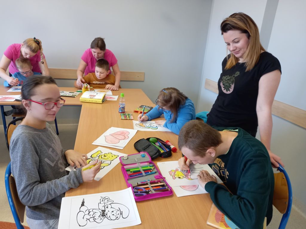 Fotografia przedstawia dzieci siedzące przy stolikach kolorujące obrazki przy pomocy opiekuna