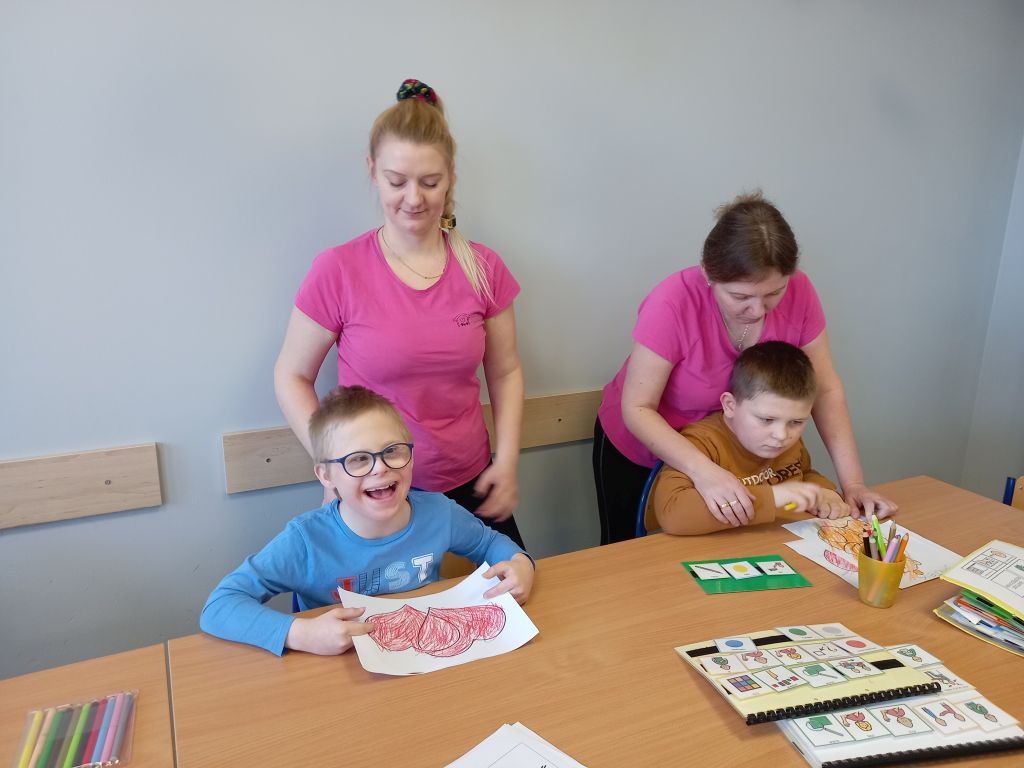 Fotografia przedstawia dzieci siedzące przy stolikach kolorujące obrazki wraz z opiekunkami