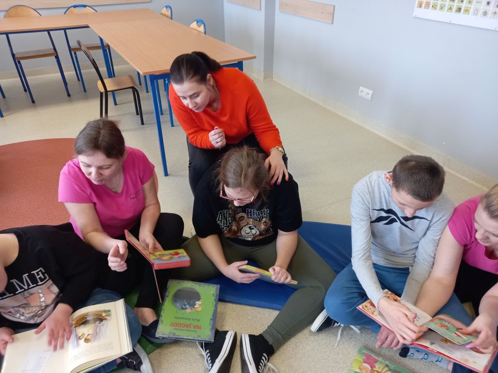 Fotografia przedstawia dzieci przeglądające kolorowe książeczki siedzące na dywanie wraz z opiekunem