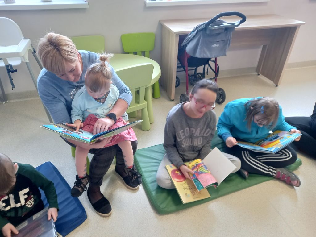 Fotografia przedstawia dzieci przeglądające kolorowe książeczki siedzące na dywanie wraz z opiekunem