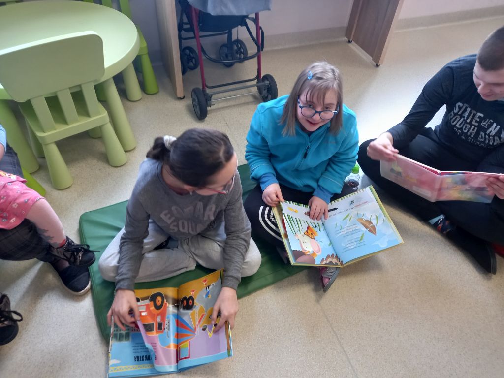 Fotografia przedstawia dzieci przeglądające kolorowe książeczki siedzące na dywanie 