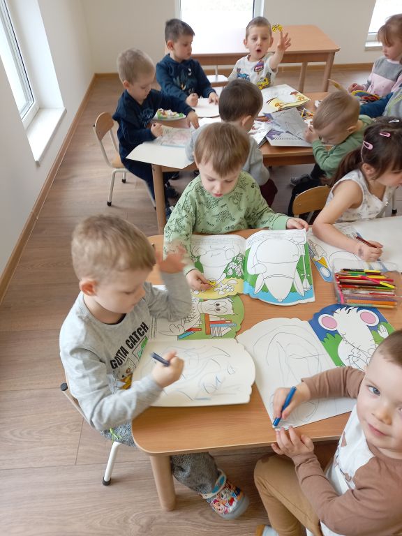 Zdjęcie przedstawia dzieci w przedszkolu siedzące przy stolikach w trakcie kolorowania książeczek