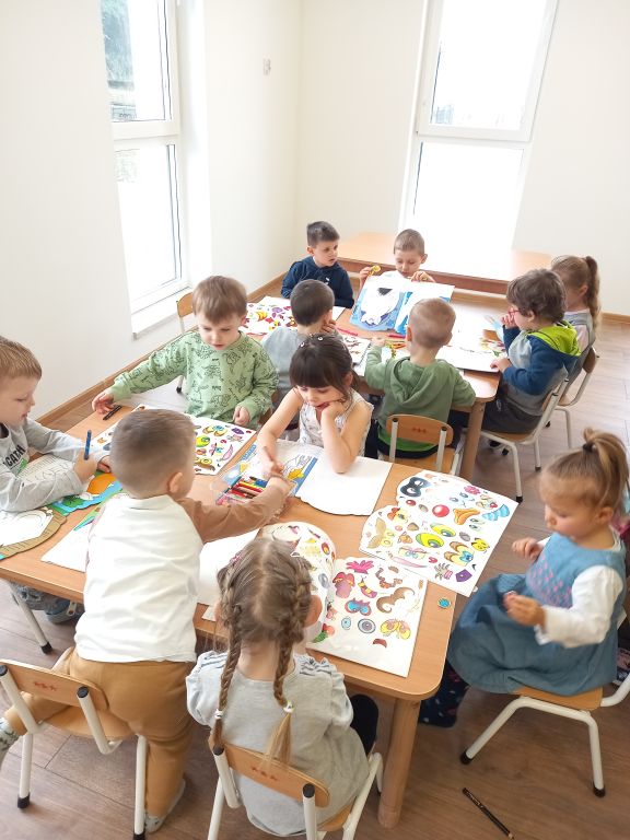 Zdjęcie przedstawia dzieci w przedszkolu siedzące przy stolikach w trakcie zajęć . Dzieci uzupełniają ilustracje w książkach naklejkami