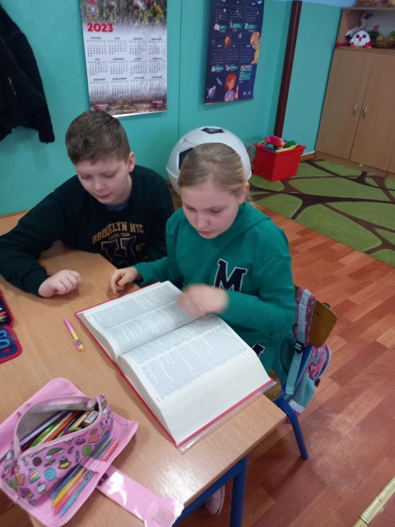 Zdjęcie przedstawia dzieci siedzące w klasie w ławkach. Dzieci przeglądają gruby słownik