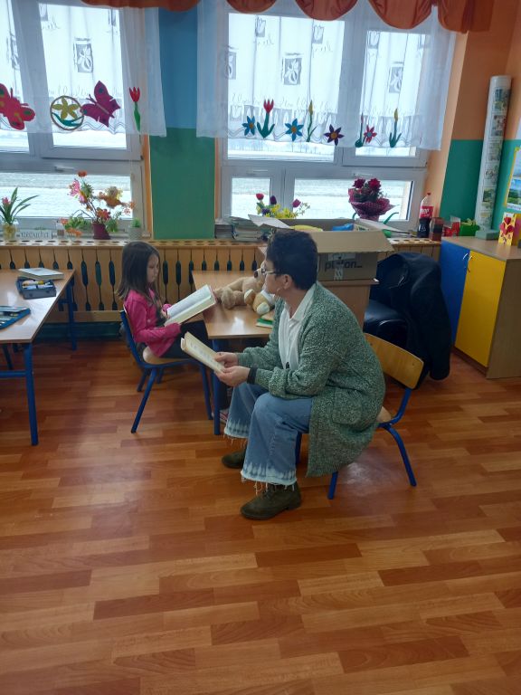 Zdjęcie przedstawia nauczycielkę siedzącą przed tablicą w klasie, czytającą dzieciom bajkę