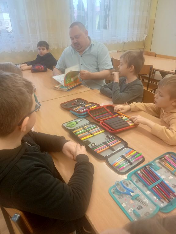 Fotografia przedstawia dzieci siedzące przy stolikach wraz z dyrektorem biblioteki, który przegląda kolorową książkę
