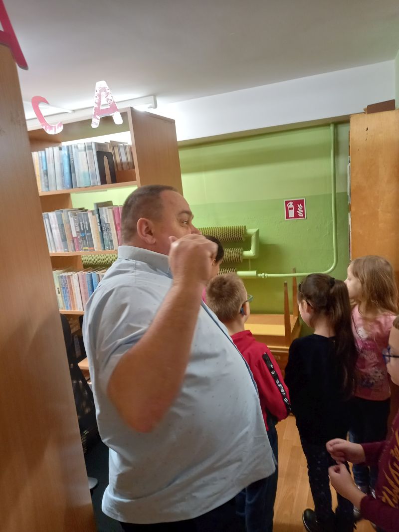 Fotografia przedstawia dyrektora biblioteki oprowadzającego dzieci wokół regałów z książkami