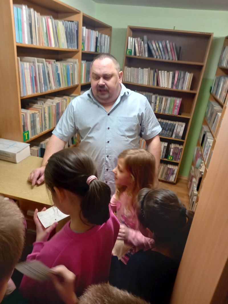 Fotografia przedstawia dyrektora pokazującego zgromadzonym wokół niego dzieciom dawny katalog biblioteczny