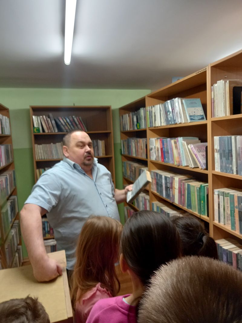 Fotografia przedstawia dyrektora biblioteki oprowadzającego dzieci wokół regałów z książkami