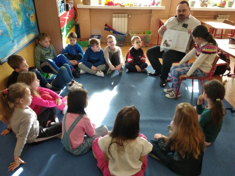 Zdjęcie przedstawia uczniów i dyrektora biblioteki siedzących w klasie na dywanie w kręgu. Dyrektor i jedna uczennica siedzą na krzesełkach. Dyrektor biblioteki trzyma w rękach książkę z ilustracjom, którą prezentuje dzieciom
