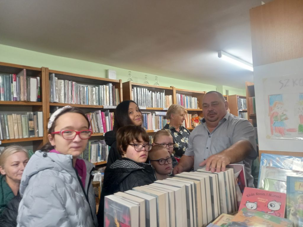 Na zdjęciu widać grupkę osób dzieci i ich opiekunów stojących w bibliotece wraz z dyrektorem biblioteki