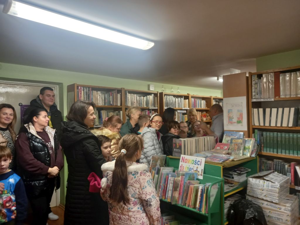 Na zdjęciu widać grupkę osób dzieci i ich opiekunów stojących w bibliotece wraz z dyrektorem biblioteki