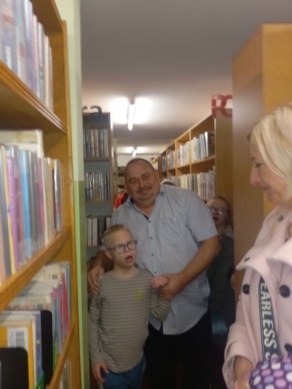 Na zdjęciu widać dyrektora biblioteki stojącego z chłopcem. W dali między regałami stoją inni uczestnicy zajęć