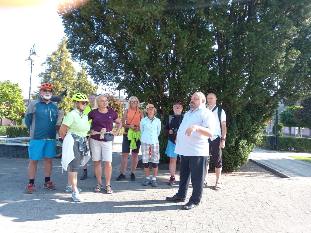Na zdjęciu widać grupę turystów i dyrektora biblioteki stojących na skwerku w przy fontannie w Skępem