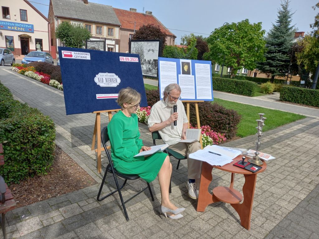 Na zdjęciu widać dwoje uczestników Narodowego Czytania siedzących na krzesłach przy stoliku na skwerku w trakcie czytania lektury