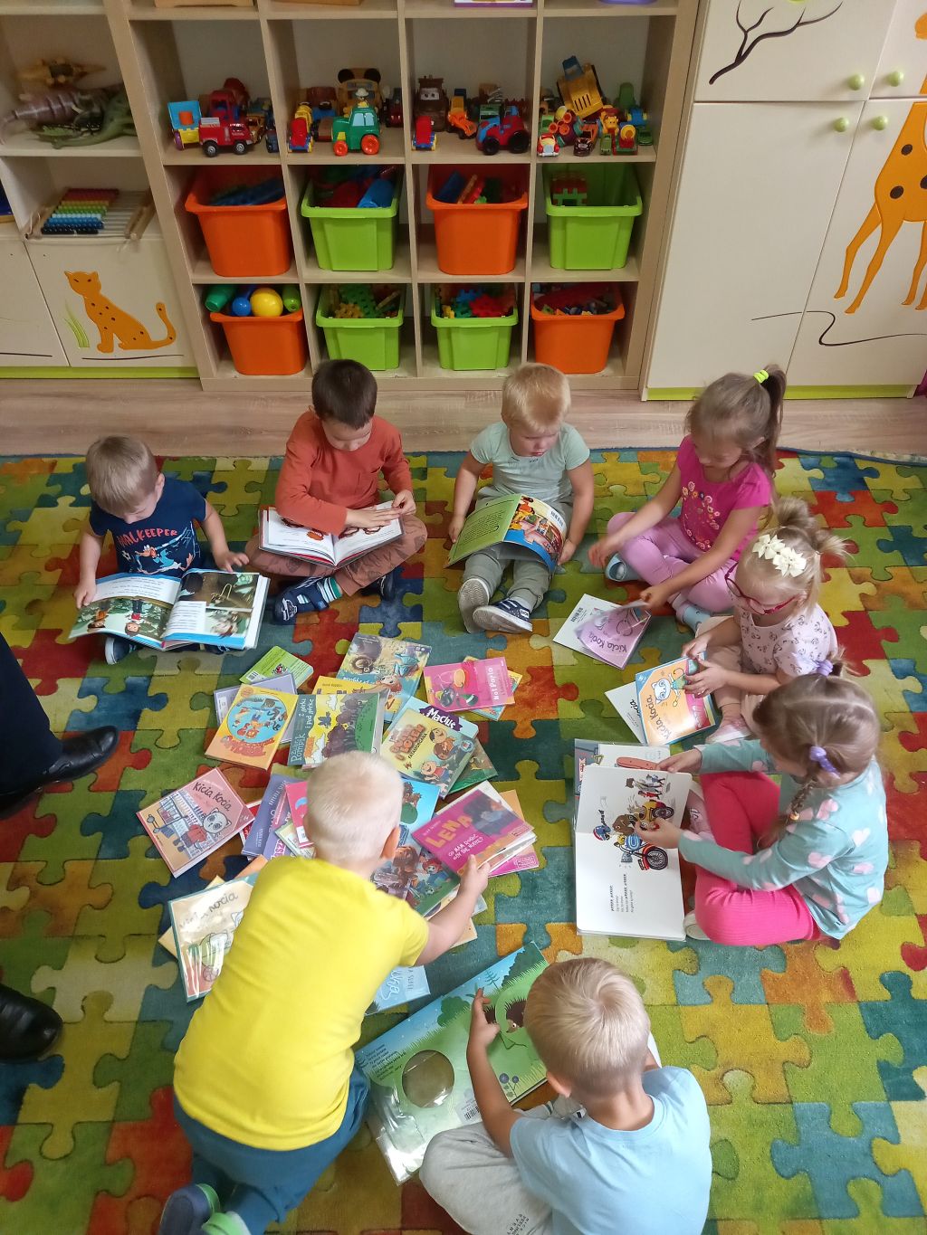 Na zdjęciu widać dzieci siedzące na dywanie w kręgu. Między dziećmi leżą książeczki, któ®e dzieci przeglądają.