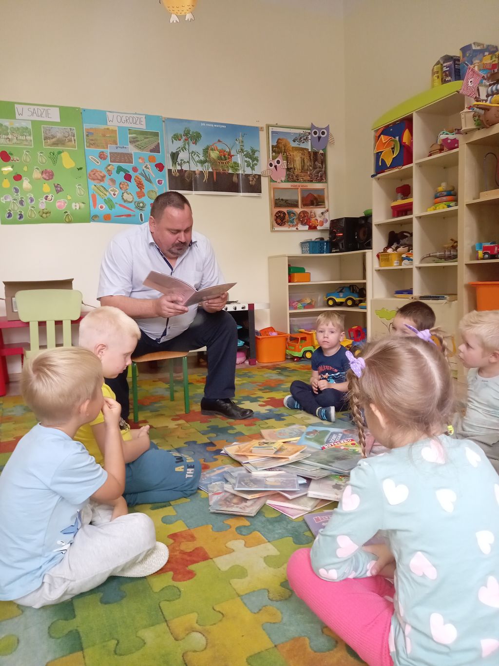 Na zdjęciu widać grupkę dzieci w siedzące na dywanie w przedszkolu. Przed dziećmi siedzi dyrektor biblioteki z książką w ręku.