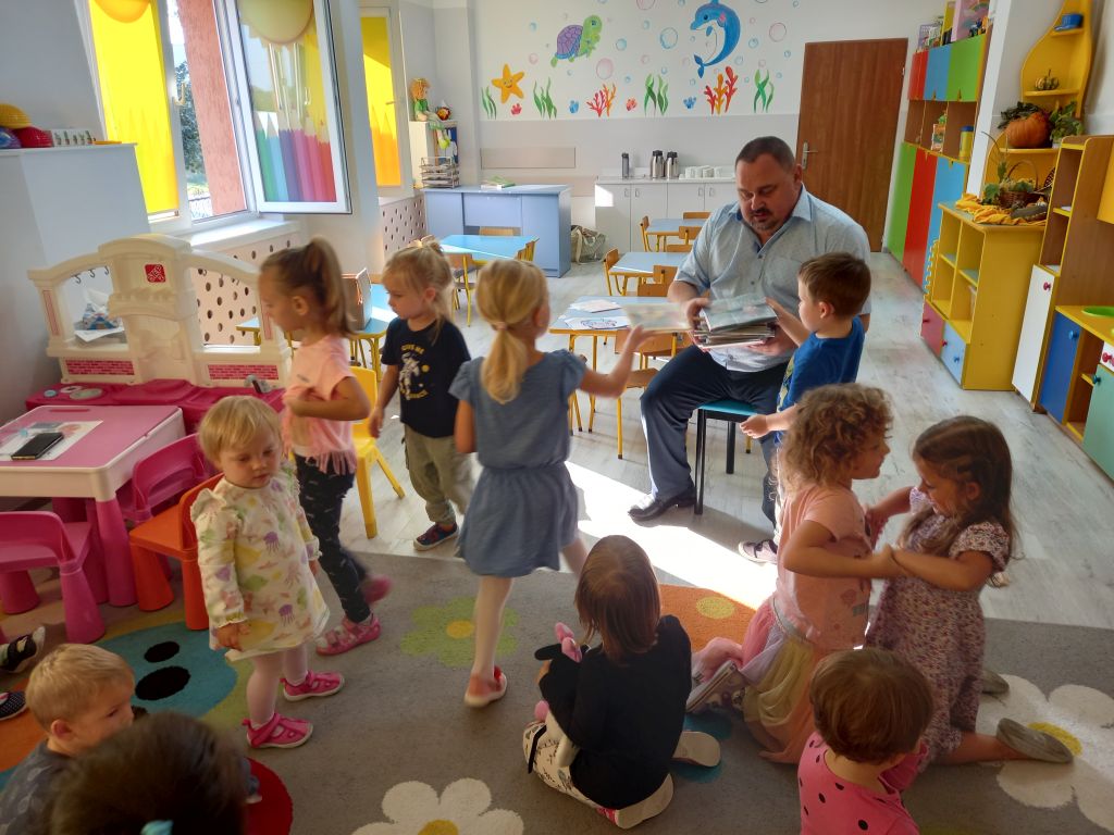 Na zdjęciu widać grupkę dzieci w przedszkolu , którym dyrektor siedzący na krześle wręcza kolorowe książecki.