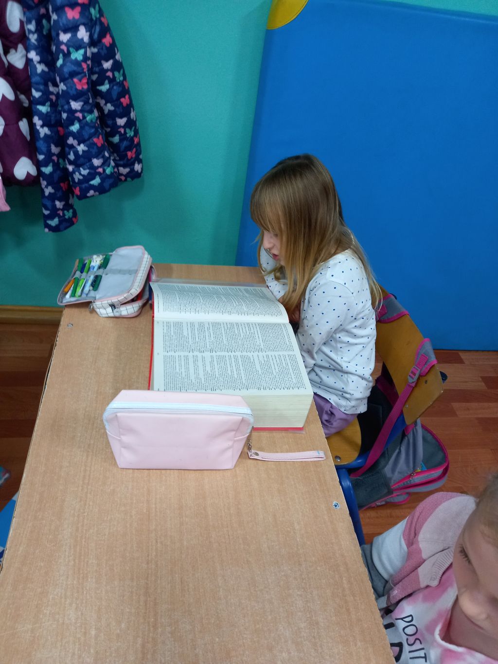 Zdjęcie przedstawia dziewczynkę siedzącą w klasie przy stoliku w trakcie czytania hasła ze słownika