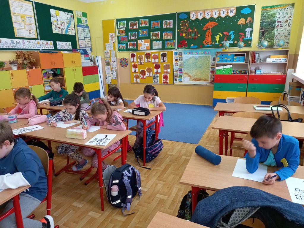 Zdjęcie przedstawia widok ogólny na dzieci siedzące w ławkach w klasie w trakcie lekcji