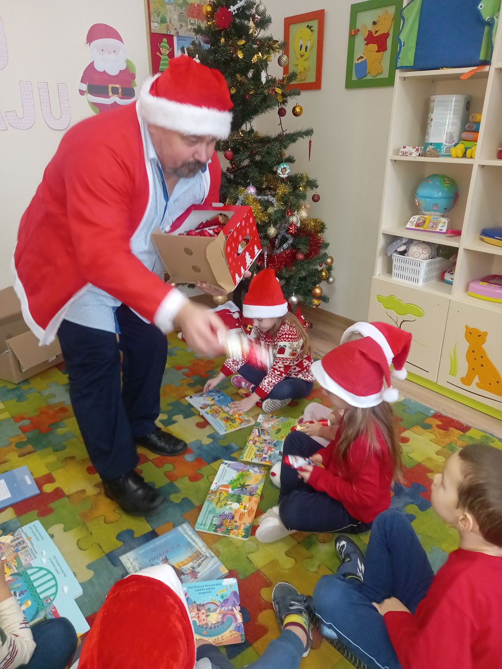 Zdjęcie przedstawia Mikołaja wręczającego dzieciom siedzącym na dywanie czekoladowe Mikołaje