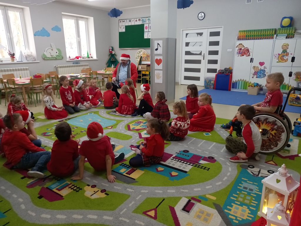 Zdjęcie przedstawia dzieci siedzące na dywanie w przedszkolu. Przed dziećmi siedzi Święty Mikołaj