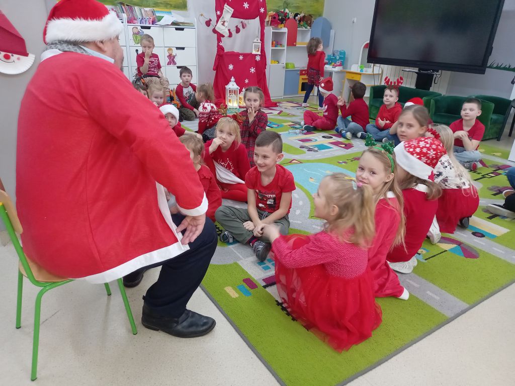 Zdjęcie przedstawia grupkę przedszkolaków siedzących na dywanie w przedszkolu przed dziećmi siedzi Mikołaj