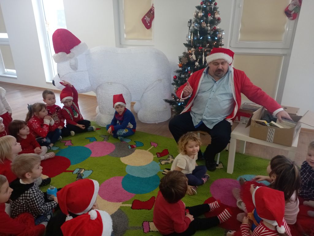Zdjęcie przedstawia grupkę przedszkolaków siedzących na dywanie w przedszkolu przed dziećmi siedzi Mikołaj oraz stoi przedszkolny opiekun