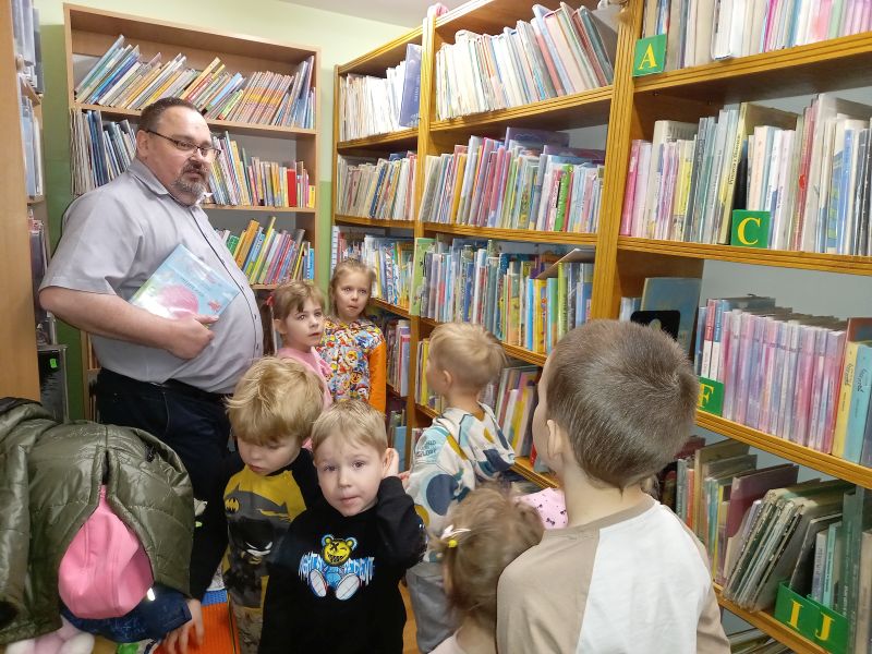 Na zdjęciu widać grupkę dzieci stojących przy regałach z książkami oraz dyrektora biblioteki oprowadzającego dzieci po bibliotece