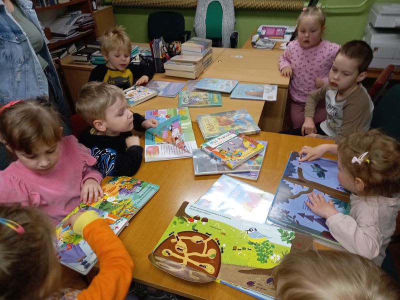 Na zdjęciu widać dzieci siedzące w bibliotece przy stolikach, na których leżą książeczki dla dzieci.