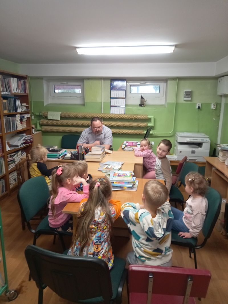 Na zdjęciu widać dzieci siedzące w bibliotece przy stolikach i dyrektora biblioteki siedzącego przy biurku przed dziećmi w trakcie prowadzonych zajęć