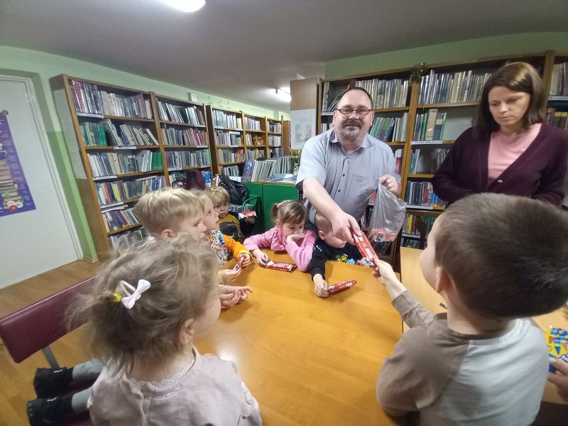 Na zdjęciu widać dzieci siedzące przy stolikach w bibliotece. Przed dziećmi stoi dyrektor i wręcza im słodycze, jako nagrodę za aktywność na zajęciach.