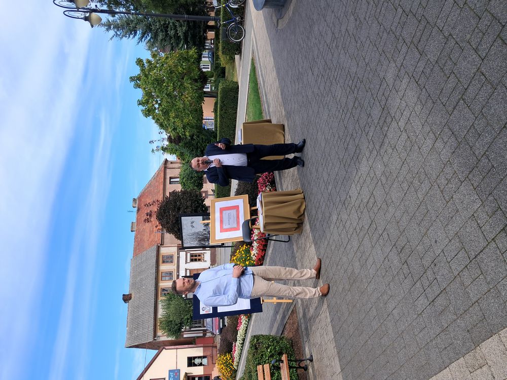 Zdjęcie przedstawia moment rozpoczęcia „Narodowego Czytania” na Rynku w Skępem. Na zdjęciu stoją Burmistrz Skępego i Dyrektor Biblioteki w Skępem. Dyrektor mówi przez mikrofon.