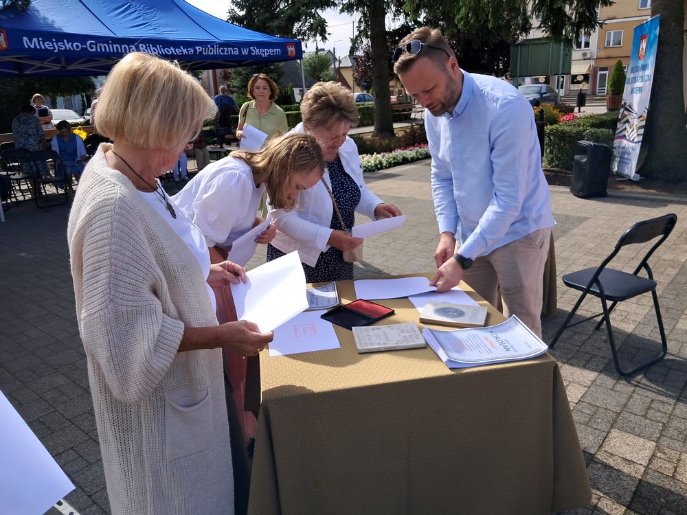 Zdjęcie przedstawia uczestników Narodowego czytania stojących przy stoliku na skwerku w Skępem. Przy stoliku stoi Burmistrz Skępego i stempluje okolicznościową pieczęcią Narodowego czytania Certyfikaty za udział w akcji.