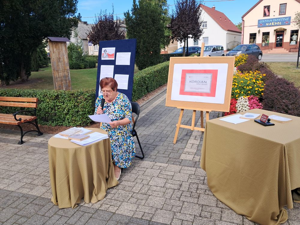 Zdjęcie przedstawia uczestniczkę Narodowego Czytania siedzącą na krześle przy stoliku na skępskim skwerku czytającą przez mikrofon lekturę 